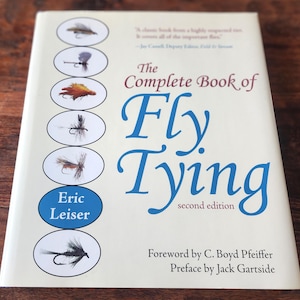 May include: A softcover book titled The Complete Book of Fly Tying, second edition, by Eric Leiser. The white cover features a vertical column of six oval illustrations depicting various fishing flies and the author's name in a blue oval.