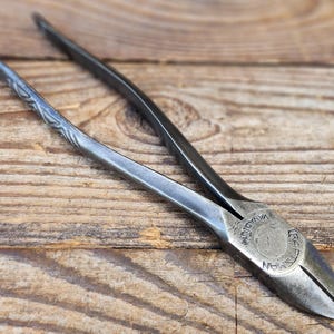May include: Close-up of a pair of Craftsman pliers, featuring a dark metallic finish. The tool rests on a wooden surface, with the brand name and "Vanadium" visible on the head. The handles have a textured design.