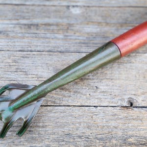 May include: A hand-held garden cultivator tool featuring a metallic green, multi-pronged head and a smooth, cylindrical red handle. The tool has a tapered neck connecting the pronged end to the handle, designed for soil aeration and weeding.