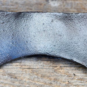 Op de afbeelding: Een dubbelzijdige bijlkop met een donkere, getextureerde, gesmede metalen afwerking. Het gereedschap heeft twee symmetrische, gebogen snijvlakken aan tegenovergestelde zijden, met een centraal oog voor de bevestiging van een steel.