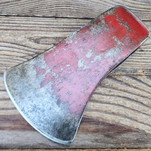 May include: A metal axe head featuring a weathered red painted finish with visible signs of wear, scratches, and exposed silver-toned metal along the curved cutting edge and the flat top surface, showcasing a rugged and aged appearance.