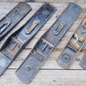 Puede incluir: Una colección de seis cuerpos de cepillos de carpintero de metal vintage dispuestos en fila, mostrando diversos grados de desgaste, oxidación y una pátina gris oscuro. Cada herramienta presenta una abertura central y nervaduras.