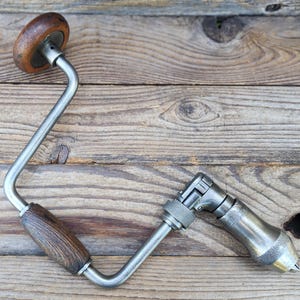 Puede incluir: Un taladro manual antiguo con mango de madera y marco de metal. El taladro tiene un diseño curvo y un portabrocas para sujetar brocas. El mango de madera es de color marrón oscuro y el metal es plateado.