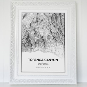 May include: A black and white topographic map print of Topanga Canyon, California. The print is framed in a white frame with a decorative border. The text "Topanga Canyon, California" and the coordinates "34&deg; 3' 57" N 118&deg; 33' 32" W" are printed below the map.