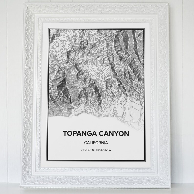 May include: A black and white topographic map print of Topanga Canyon, California. The print is framed in a white frame with a decorative border. The text "Topanga Canyon, California" and the coordinates "34&deg; 3' 57" N 118&deg; 33' 32" W" are printed below the map.