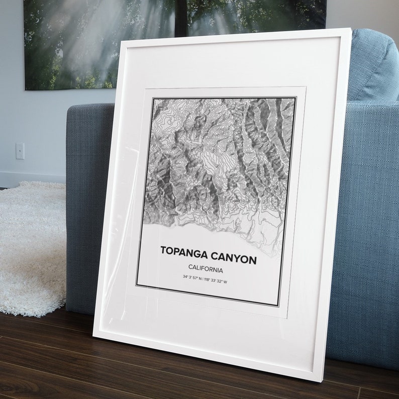 May include: A white framed print of a topographic map of Topanga Canyon, California. The map shows the elevation contours of the canyon. The text "Topanga Canyon, California, 34&deg; 3' 57" N 118&deg; 33' 29" W" is printed below the map.