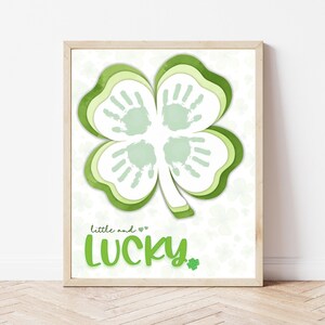 Puede incluir: Un trébol de cuatro hojas verde con huellas de manos en el centro. El trébol está rodeado de un fondo blanco con pequeños tréboles verdes. El texto "little and ♥ lucky" está debajo del trébol.