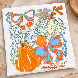 May include: A white fabric square featuring a colorful graphic print of two pumpkins, three decorative bows, and several flowers. The design uses bright orange, light blue, and cream colors with gold glitter outlines on all the illustrated elements.