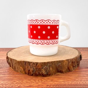 Op de afbeelding: Witte keramische mok met een rood-wit stippenpatroon en een witte kantversiering rond de bovenkant. De mok staat op een houten schijf.