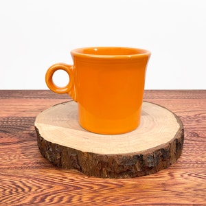Puede incluir: Una taza de cerámica de color naranja brillante con un asa redondeada. La taza está sobre una posavasos de madera.