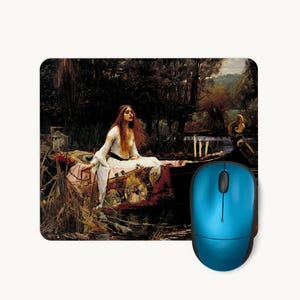 May include: A rectangular mouse pad featuring a reproduction of the painting The Lady of Shalott, showing a woman with long red hair in a white dress sitting in a boat, paired with a solid bright blue computer mouse with a black scroll wheel.