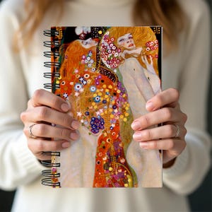 May include: Spiral-bound notebook with a colourful artistic print. The cover displays a detailed painting with figures, intricate floral patterns, and a warm palette of reds, oranges, and blues. The notebook's spiral binding is black.