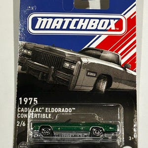 Op de afbeelding: Een groene 1975 Cadillac Eldorado cabriolet Matchbox speelgoedauto. De verpakking heeft een blauw en rood ontwerp met het Matchbox-logo en de tekst "Designed for a Lifetime".