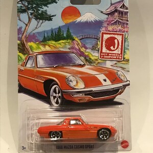 May include: A red 1968 Mazda Cosmo Sport Hot Wheels die-cast car on a blue and white card with a Japanese mountain scene. The card also features the Hot Wheels J-Imports logo.