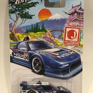 Puede incluir: Un modelo de coche de fundición a presión Hot Wheels J-Imports azul y blanco de un Acura NSX con el número 24 en el lateral. El coche está en una tarjeta blanca con un fondo azul y una imagen de una escena de montaña japonesa.