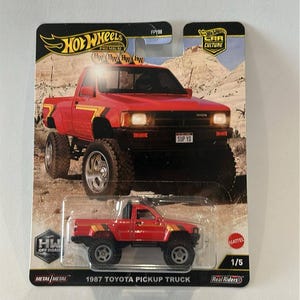 May include: A red 1987 Toyota pickup truck toy with black detailing and large off-road tyres. The packaging includes the text "Hot Wheels Premium" and "Car Culture". The text "SUP YO" is visible on the truck.