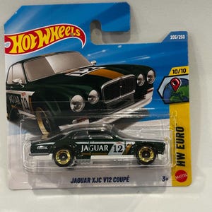 Może przedstawiać: Model samochodu Hot Wheels Jaguar XJC V12 Coupé w kolorze ciemnozielonym z białymi i złotymi pasami wyścigowymi, numerem 12 oraz logo Castrol. Samochód znajduje się w oryginalnym niebiesko-żółtym opakowaniu typu blister z napisem HW EURO.