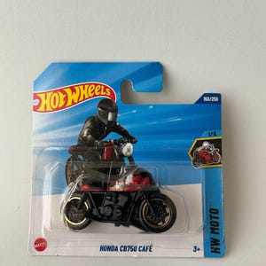 Op de afbeelding: Een Hot Wheels Honda CB750 Café motorfiets speelgoed in een doorzichtige plastic verpakking. De motorfiets is zwart en rood met goudkleurige accenten. De verpakking heeft een blauw en wit ontwerp met het Hot Wheels logo en de tekst "KW Moto".
