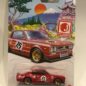 Op de afbeelding: Rode Nissan Skyline HT 2000GT-X Hot Wheels die-cast auto met gouden wielen en "15" aan de zijkant. De auto staat op een witte achtergrond met een Japans berglandschap en de tekst "Hot Wheels J-Imports".