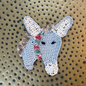 May include: A gray crocheted donkey with a white and brown mane. The donkey has a red, green, and white striped mane and a white nose.