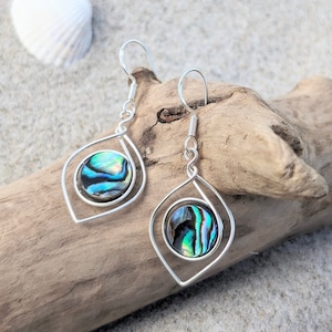 Könnte beinhalten: Ein Paar silberne Ohrringe mit einem tropfenförmigen Design. Jeder Ohrring hat eine runde Abalone-Muschel mit schillernden grünen, blauen und violetten Farben. Die Muschel ist in einem silbernen Drahtrahmen gefasst.