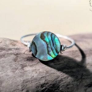 Könnte beinhalten: Ein silberner Ring mit einem runden Einlegeteil aus Abalone-Muschel. Die Muschel hat ein blaues, grünes und braunes irisierendes Muster.