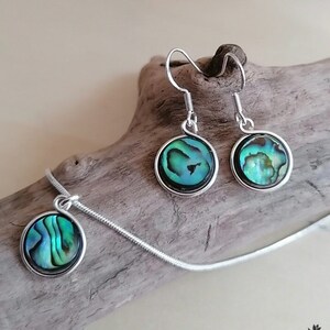 Könnte beinhalten: Ein silbernes Halsketten- und Ohrring-Set mit schillernden grünen Abalone-Muschel-Anhängern. Die Halskette hat eine zarte Kette und die Ohrringe haben einfache silberne Haken.