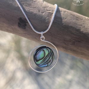 Könnte beinhalten: Eine silberne Halskette mit einem Anhänger in Spiralform. Der Anhänger enthält eine runde, irisierende Abalone-Schale. Die Schale zeigt Farbtöne von Blau, Grün und Lila. Die Halskette wird auf einem Stück Treibholz präsentiert.