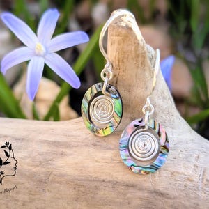 Könnte beinhalten: Ein Paar silberne Ohrringe mit einem Spiraldesign und irisierenden Abalone-Muschelakzenten. Die Ohrringe hängen an einer Holzoberfläche.