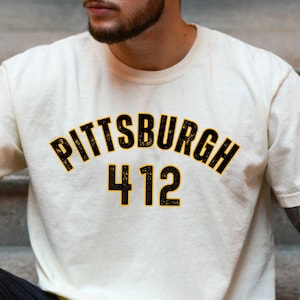 May include: A cream colored t-shirt with a black and yellow graphic that says "Pittsburgh 412".