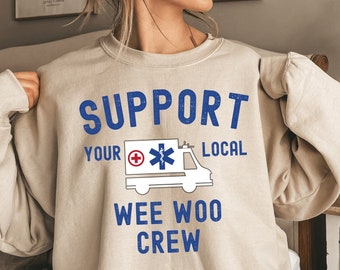 以下が含まれることがあります： 青い文字で「SUPPORT YOUR LOCAL WEE WOO CREW」と書かれたベージュのトレーナー。テキストの隣には、赤い十字架と青い生命の星が描かれた白い救急車が描かれています。