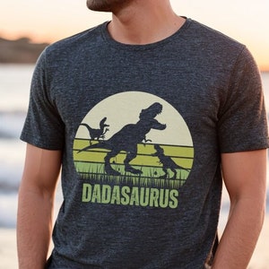 May include: Dark gray t-shirt featuring a dinosaur graphic with the text "DADASAURUS". The design includes a silhouette of a T-Rex and smaller dinosaurs, set against a yellow and green background resembling a sunset.