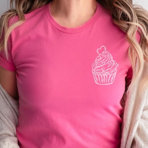 May include: A pink t-shirt with a white outline of a cupcake with hearts on it.