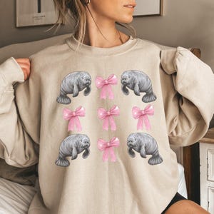 May include: Beige sweatshirt featuring a pattern of gray manatees and pink bows. The manatees are depicted in a realistic style, while the bows are a bright pink color. The design is repeated across the front of the sweatshirt.