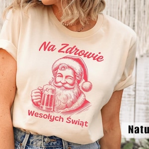May include: A natural-colored t-shirt featuring a red illustration of Santa Claus holding a beer stein. The text "Na Zdrowie" and "Wesołych Świąt" are printed above and below the Santa graphic, respectively. The shirt has short sleeves and a crew neck.