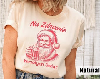 以下が含まれることがあります： ビールジョッキを持ったサンタクロースの赤いイラストがプリントされた、ナチュラルカラーのTシャツ。「Na Zdrowie」と「Wesołych Świąt」の文字がサンタのグラフィックの上と下にプリントされています。半袖でクルーネックのシャツです。