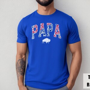 May include: A true royal blue short-sleeve t-shirt featuring the word PAPA in a red, white, and blue zebra-striped pattern across the chest. Below the text is a solid white silhouette of a buffalo. The shirt has a crew neckline and a standard fit.