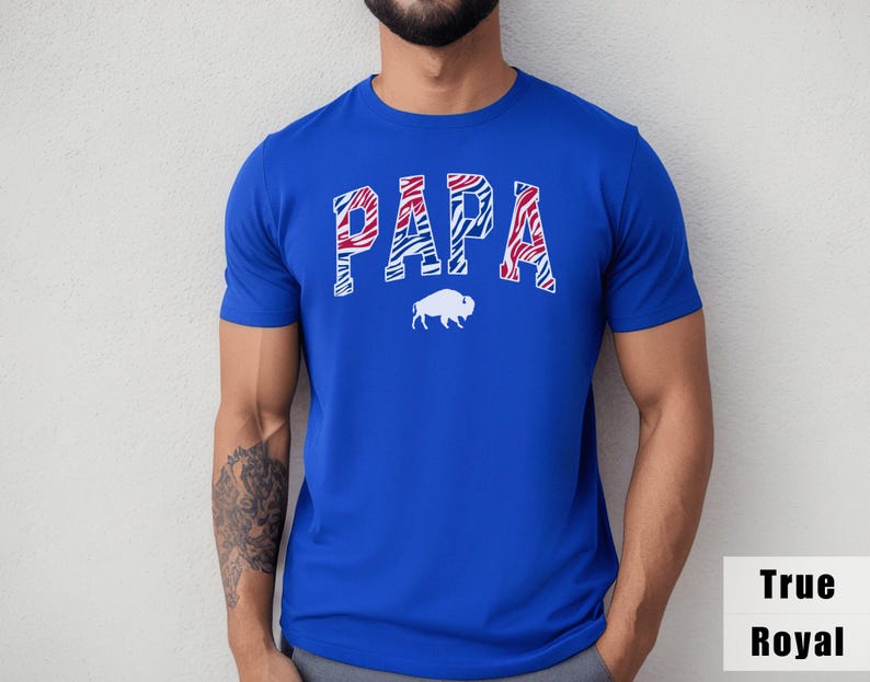 May include: A true royal blue short-sleeve t-shirt featuring the word PAPA in a red, white, and blue zebra-striped pattern across the chest. Below the text is a solid white silhouette of a buffalo. The shirt has a crew neckline and a standard fit.