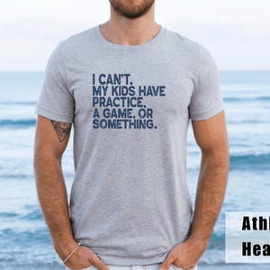 May include: Athletic heather gray t-shirt with the navy blue text: "I CAN'T. MY KIDS HAVE PRACTICE, A GAME, OR SOMETHING." The shirt is a crew neck style and is made of a soft, comfortable material.