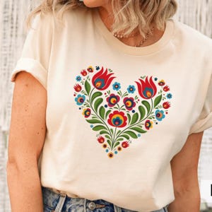 May include: Cream-colored t-shirt featuring a heart-shaped floral design. The heart is composed of red tulips, blue and yellow flowers, and green leaves. The shirt has short sleeves and a crew neck.