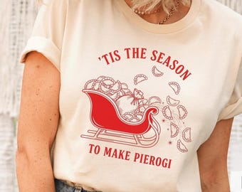 May include: Cream-colored t-shirt with rolled sleeves featuring a red illustration of a sleigh filled with pierogi. The text above reads, "'Tis the Season" and below, "To Make Pierogi."