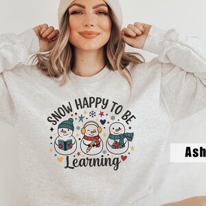 May include: Ash gray sweatshirt with a graphic of three snowmen wearing hats, scarves, and earmuffs. The text "SNOW HAPPY TO BE Learning" is printed above the snowmen in a circular design. The snowmen are holding a book, a pencil, and an apple.