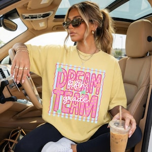 May include: A pale yellow short-sleeved t-shirt featuring a graphic print with the text "DREAM TEAM" in large pink polka-dot letters and "fifth grade" in white script. The design includes light blue stripes and small green stars.