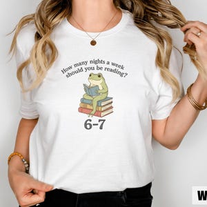 May include: White t-shirt with a whimsical graphic of a green frog reading on a stack of books. The text reads, "How many nights a week should you be reading?" and "6-7". A fun, literary-themed tee.