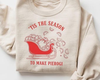 May include: Beige sweatshirt with a red graphic of a sleigh filled with pierogi and the text "'Tis the Season to Make Pierogi". The design includes falling pierogi and a star.