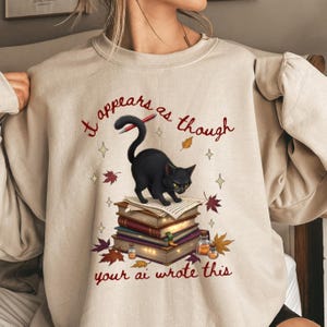 May include: Beige sweatshirt with a black cat illustration standing on a stack of books. The text "It appears as though your ai wrote this" is printed in red. Autumn leaves and stars surround the cat.