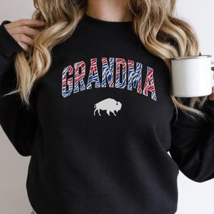 May include: A black crewneck sweatshirt featuring the word GRANDMA in an arched, red and blue zebra-striped pattern. Below the text is a solid white silhouette of a buffalo. The garment has long sleeves and a ribbed neckline.