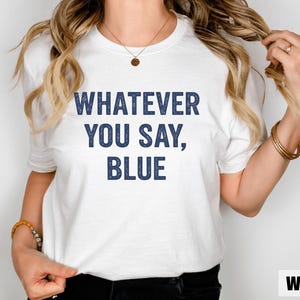 May include: A white t-shirt with the text "WHATEVER YOU SAY, BLUE" in navy blue block letters. The shirt is a crew neck style and is being worn by a person. The word "White" is in the bottom right corner.