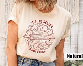 May include: A natural-colored t-shirt with a burgundy graphic. The graphic features a rolling pin surrounded by pierogi and the text "'Tis the season to make pierogi."