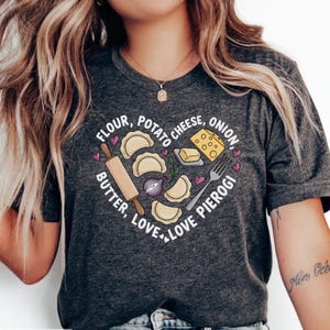 May include: Dark gray t-shirt with a heart-shaped design featuring pierogi ingredients: flour, potato, cheese, onion, butter, and love. The design includes a rolling pin, fork, and pierogi illustrations.